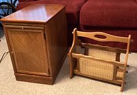 Side table with cabinet door and cane wood magazine rack placed on carpet flooring in front of a red couch.
