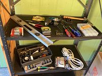 Photo showing a variety of tools and hardware items on a black shelving unit including a long metal level, tape measure, hammer, electric drill with bits, screwdrivers, pliers, utility knife, small hardware organizer box, and extension cord.