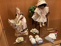 Wide shot showing vintage porcelain lady leaning on floral vase, seated porcelain doll in lace dress and pink hat, two porcelain dolls, porcelain roses, and miniature gardening tools.