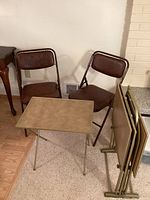 Photo showing two brown folding chairs with metal frames and one rectangular vintage folding TV tray fully opened next to a stand holding three TV trays in a folded position.