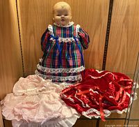 Vintage baby doll wearing plaid dress with lace, alongside pink and red dresses laid out as alternative outfits, wood panel background.