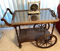 Full view of the vintage wooden tea cart with removable glass tray top and large spoked wheels, with brass and glass desk clock placed on top.