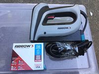 Electric gray and black Arrow staple tool with attached power cord placed flat on a surface next to a box of Arrow T50 staples, showing brand names and product details