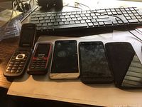 Five cell phones displayed in a row on a desk in front of a keyboard, including two flip phones (one Samsung), a cracked screen Samsung smartphone, and other smartphones.