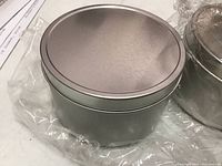 Photo of one deep silver metal tin with lid on a surface, partially wrapped in clear plastic.