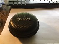 Black round zippered carrying case with Oranka branding on top, textured surface.