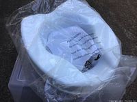 White raised toilet seat in original clear plastic packaging with visible warning labels.