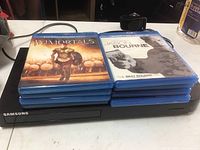 Samsung black Blu-Ray player with 8 Blu-Ray movies stacked on top, including Immortals and The Best Bourne.