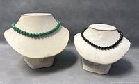 Two beaded jade necklaces displayed on white bust stands: a green beaded choker and a black beaded longer necklace.
