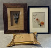 Three framed pieces shown, front view: carved wooden mask artwork, rabbit sketch with flowers, and empty ornate gold frame lying flat