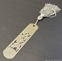 Close-up image of the silver bookmark showing its floral cut-out design and attached tassel on a dark backdrop.