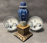 Full view of the lot showing two 8-inch Chinese porcelain plates with scenic black and red designs, a tall blue and white porcelain jar, a small floral patterned toothpick holder, and a square wooden base with Chinese calligraphy.