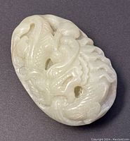 Front and angle views of an oval-shaped pale green jade pendant with a detailed carved dragon motif covering the surface.