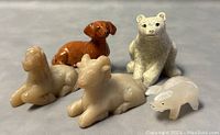 Five small animal figurines on a gray surface including a brown Dachshund, beige bear, cow, horse, and a small onyx pig.