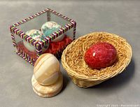 View of two hand-painted Chinese eggs in decorative glass display case alongside red stone egg in straw basket and polished marble egg on stand.