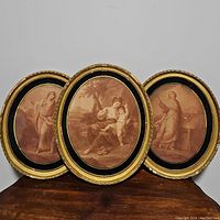 Three oval framed sepia tone art prints with classical figures in gold and black oval frames on a wooden surface
