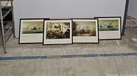 Four framed battle art prints of naval battles placed on the floor in a row.