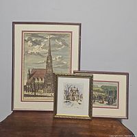 Three framed artworks arranged on a wooden surface against a wall, showing a Gothic cathedral print, a winter scene, and a crowd scene, with visible frame wear.
