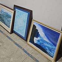 Photo shows three framed pieces leaning against a wall on a concrete floor: two paintings and one map frame in the middle with loose frame edge on the map.