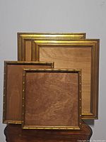 Four wooden picture frames with gold finish, various sizes, empty with wooden backings visible, simple classic design
