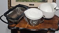 Photo showing glass measuring cup with white lid and strainer inside, black mesh strainer with handles, and three white mixing bowls.