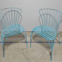 Pair of blue metal outdoor chairs shown side by side on cement floor against a white backdrop, facing forward to display front and backrest shape and seat design.