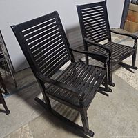 Two black outdoor wooden rocking chairs side by side on a concrete floor, showing the overall size and style.