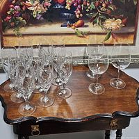 Group of 12 smaller stem flute glasses with slender stems and decorative engraving, shown together with 6 larger clear wine glasses on a wooden table.