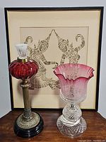 Full view of vintage red glass lamp with ornate base and pink candle holder with etched floral design, set against a framed artwork.