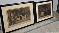Two black framed vintage prints of groups of people gathered outdoors, standing on floor against wall.