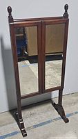 Full view of vintage cheval mirror standing upright, showing mirrored doors and wooden frame with finials and curved legs.