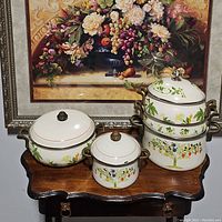 Five pieces vintage floral enamel cookware set displayed on wood table with painting backdrop.