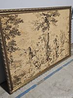 Full view of the large vintage tapestry in its carved gold frame showing the entire artwork with figures, trees and horses in a beige monochrome palette.