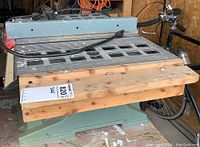 Side view showing greenish metal base, plastic/metal tabletop with grid design, attached wooden plank, and attached blade underneath.