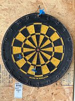 Wall-mounted black and yellow dartboard with two blue darts inserted, mounted on wood with visible wear.