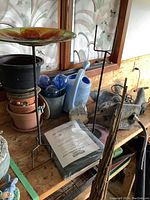 Photo showing stacked planters, colorful glass watering stakes in a metal bucket, blue watering can, concrete fountain fixture, bamboo plant trellis sticks, and decorative bird baths on stands