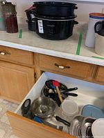 Photo of black Crock Pot slow cooker and assorted kitchen items including pots, lids, utensils in a lower cabinet drawer