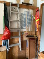 Full view of aluminum ladder mounted on wall with brooms, car creeper, and safety vest surrounding it