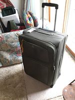 Large black rolling suitcase standing upright with wheels and a telescoping handle, near a floral fabric item and glass door.