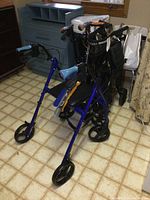 Blue folding wheeled walker with brakes and black seat. Shows part of the background furniture.