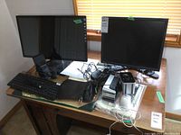 Two monitors on a table with keyboard, mouse, speakers, and cords tangled on the surface