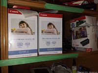 Two boxed Canon Selphy CP photo printers shown side by side with paper and ink sets. One box unopened, the other opened with receipt attached.