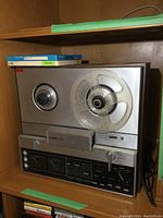 Front view of Ampex AX-50 reel to reel tape player with one clear reel loaded and two boxed spare reels on shelf above.