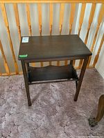Front view of a dark stained solid wood side table with a lower magazine shelf and four straight legs, placed on a carpeted floor near wooden balusters.