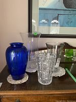 Photo showing four vases placed on a wooden surface under a framed artwork. The blue glass vase is on the left, the clear crystal vase with patterns is front center, and two smooth clear glass vases with flared tops are at back right.