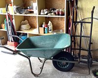 Green plastic wheelbarrow with metal frame and black grip handles in a garage setting, showing general condition.