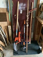 Full view of yard tools including shovel, hedge trimmer, pruner, rake, pitchfork, and weed puller arranged against wall on a dark mat.