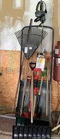 View of multiple yard tools including several snow shovels, garden shovels, and a large fan rake leaning against a metal frame in a garage.