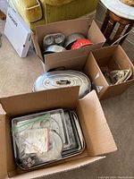 Wider view of boxes containing various metal baking pans, muffin pans, and a large covered turkey roaster with metal handle.