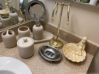 Photo of bathroom counter showing ceramic soap dispenser, toothbrush holder, covered jar, tray, silver soap dish, brass towel hanger, ceramic mermaid dish, and magnifying mirror